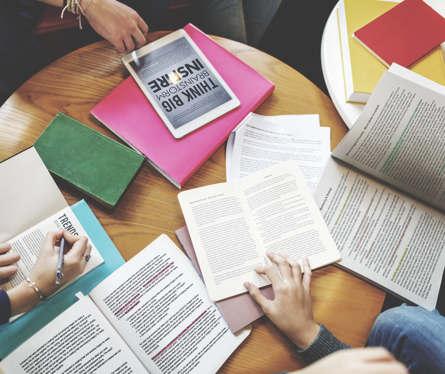 BD Medical Writing & Consulting - Draufsicht auf einen Holzschreibtisch: Offene Bücher, ein Tablet und verstreute Papiere liegen herum. Eine Hand hält ein Buch offen.